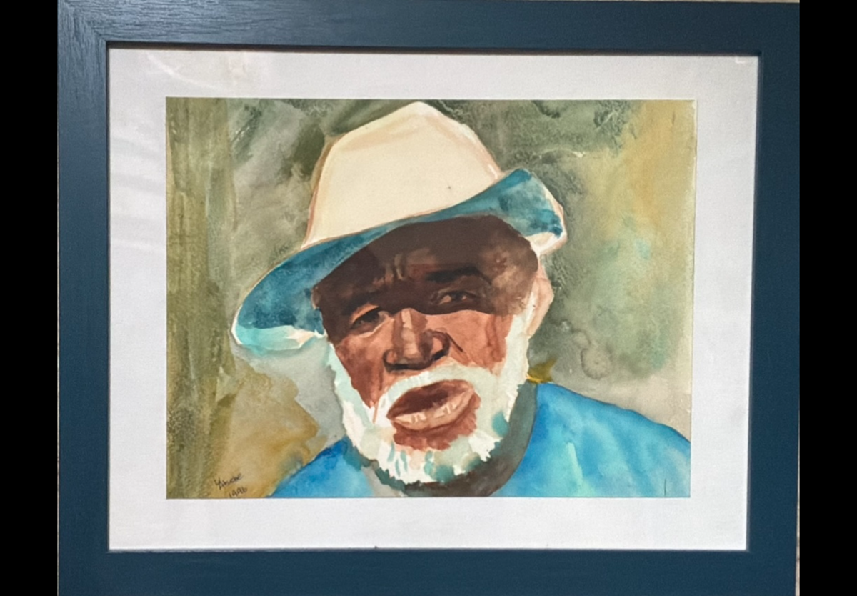 A framed watercolor portrait painting of an elderly man wearing a light-colored hat and blue shirt, set within a dark blue frame and white mat.